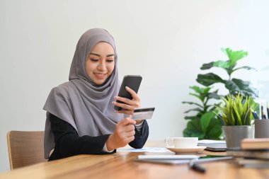 Hoş Asyalı Müslüman kadın kredi kartı tutuyor ve online alışveriş için akıllı telefon kullanıyor..