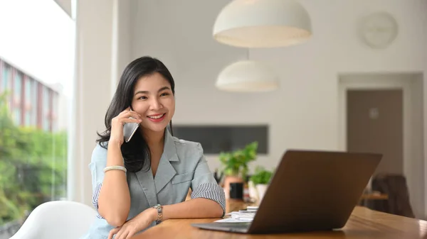 漂亮的年轻亚洲女人 电话交谈愉快 在现代办公室使用笔记本电脑 — 图库照片