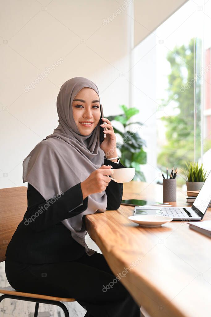 Smiling Muslim woman in hijab having pleasant phone conversation and drinking coffee at her workplace.