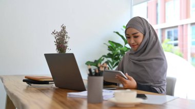 Beautiful muslim woman sitting front of laptop and using digital tablet at home office.