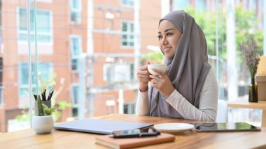 Düşünceli genç Müslüman kadın elinde kahve fincanı tutarak pencereden dışarı bakıyor, yeni başarılı projelerin hayalini kuruyor..