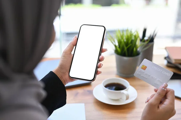 Over shoulder view of Muslim woman holding credit card and using mobile phone. Online shopping, internet banking concept.