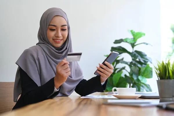 Smiling muslim woman holding credit card and using smart phone, purchasing product via online shopping.
