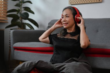 Carefree middle aged woman wearing wireless headphones, listening to music, relaxing on sofa at home.