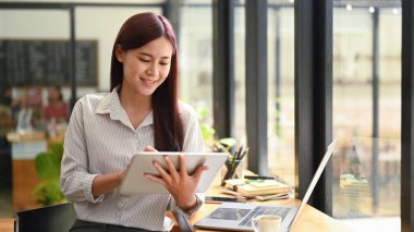 Gülümseyen genç kadın ekonomist modern kahve dükkanında otururken dijital tablette finansal verileri kontrol ediyor..