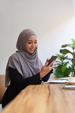 Gülümseyen Müslüman kadın sosyal medyayı kontrol etmek ya da çevrimiçi iletişim kurmak için cep telefonu kullanıyor.