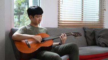 Genç adam gündelik kıyafetlerle kanepede akustik gitar çalıyor, evde kaygısızca vakit geçiriyor..