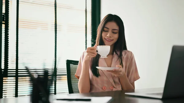 Happy young woman entrepreneur enjoying her morning coffee, sitting in contemporary office.