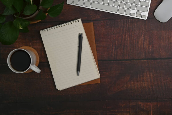 Blank notepad, coffee cup, wireless keyboard and houseplant on wooden table.
