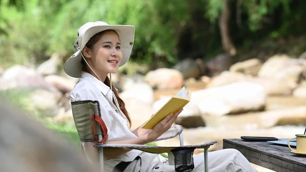 Peaceful female traveler resting in folding chair near the river bank. Adventure, travel, tourism and camping concept.