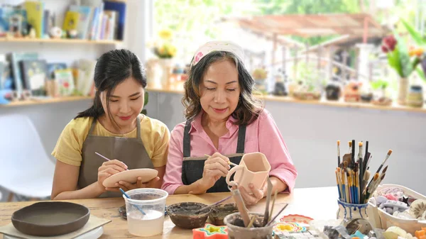 Young woman and retired woman painting painting on bowl before baking in pottery workshop.