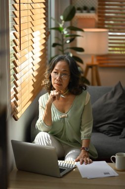 Orta yaşlı bir kadın kanepede dinleniyor ve pencereden bakıyor. Stressiz ve huzurlu bir evde keyfine bakıyor..