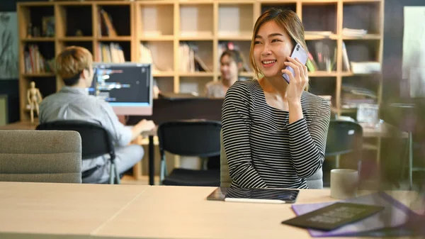 Cheerful Creative Woman Talking Mobile Phone Her Colleague Sitting Background — Φωτογραφία Αρχείου