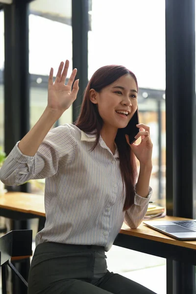 Cheerful female employee waving hands to greet her colleagues while talking on mobile phone.