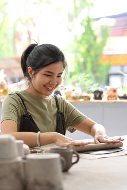 Gülümseyen kadın kil kütle için merdane kullanıyor ve bir masada seramik yapıyor, atölyede el yapımı seramik yapıyor..