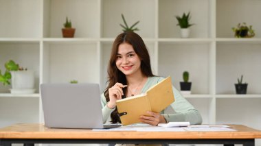 Güzel, genç ve serbest çalışan bir bayan modern ofiste oturuyor ve kameraya gülümsüyor..