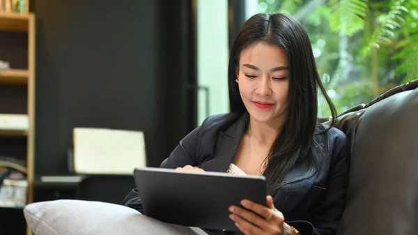 Beautiful millennial businesswoman resting on couch at her personal office and using digital tablet.