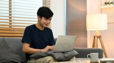 Smiling young asian man relaxing on couch and using laptop computer.