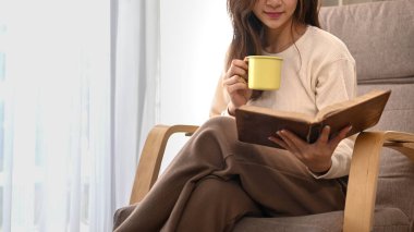 Smiling young woman drinking herbal tea and reading book on armchair, spending leisure weekend at home.