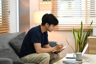 Side view asian male freelancer working online with laptop computer on couch at home.