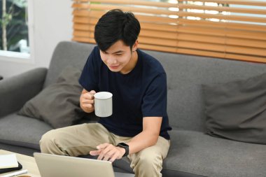 Asian man in casual clothes drinking coffee and surfing internet or checking email on laptop at home.