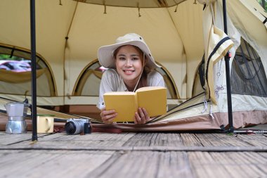 Cheerful young woman lying in her camping tent and reading a book. Travel, camping and vacation concept.