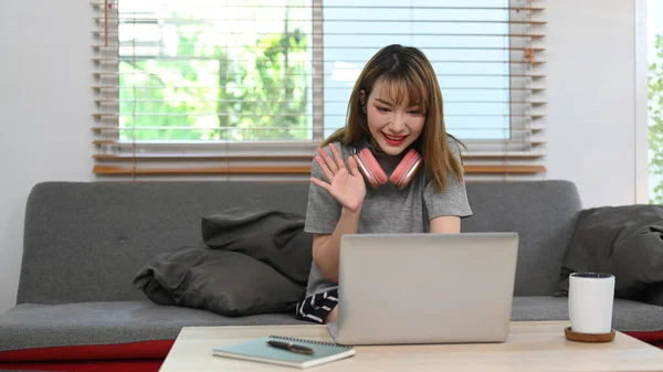 Smiling young woman chatting online, making video call via laptop while sitting on couch at home.