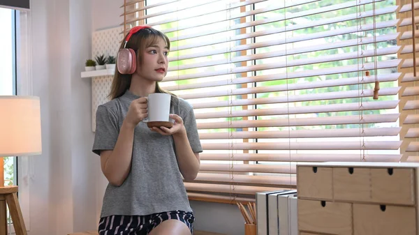 Satisfied young woman holding coffee cup and looking through the window, enjoy stress free peaceful mood.