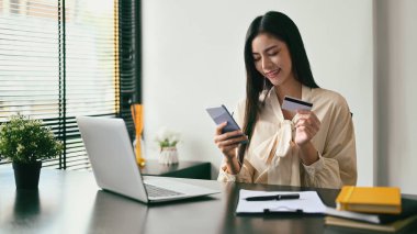 Smiling young female holding credit card, making payment online on her mobile phone.