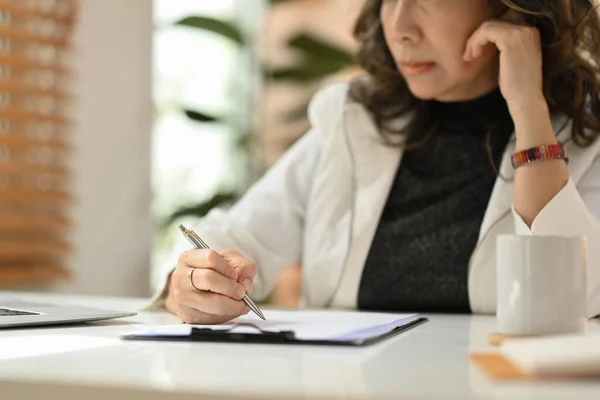 Focused mature woman remote working from home, writing information on notebook.