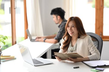 Cheerful female employee holding notebook and using laptop at her office desk with her colleague.