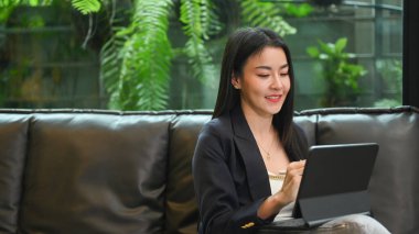 Charming millennial working woman resting on couch at her personal office and using digital tablet.