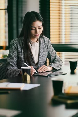 Concentrated female manager sitting at her workplace and writing important information on notebook.
