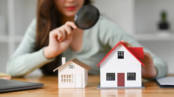 Woman looking at house models through magnifying glass. House selection, real estate appraisal and house search concept.