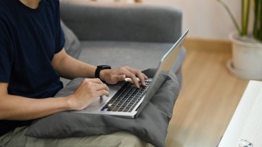Cropped view of man sitting on couch and working online or surfing internet with laptop.