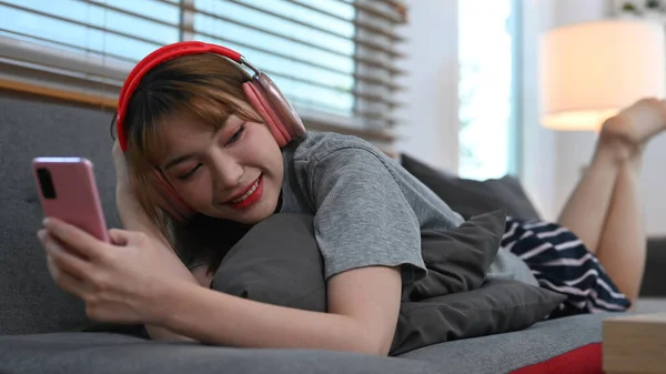 Happy Young Woman Wearing Headphone Lying Couch Using Mobile Phone — 스톡 사진
