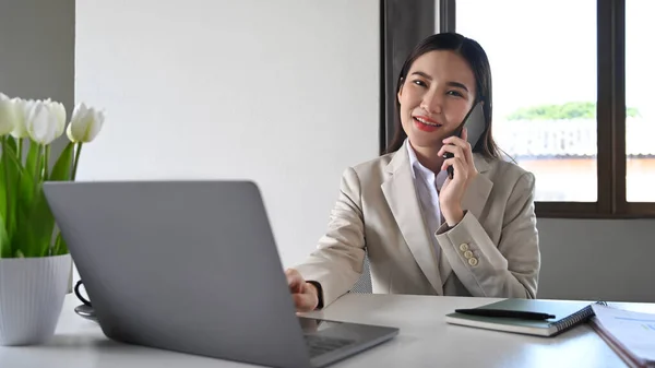 Gorgeous asian woman in business suit talking on mobile phone and using laptop at her office.