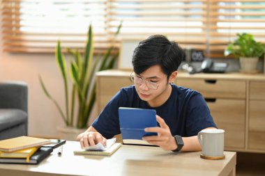 Focused asian man calculating household finances or taxes on calculator, sitting in bright living room.