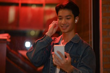 Smiling hipster man standing under neon light in night city street and using smart phone.