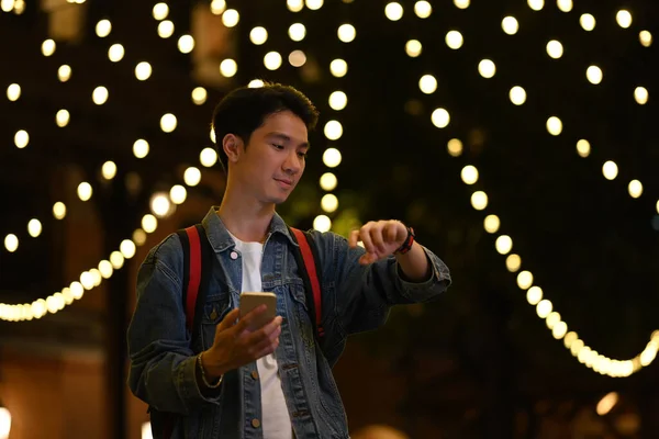 Young man standing in city avenue at night and typing text message on smart phone.