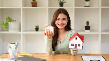 Pretty young woman holding house models and smiling at camera. Real estate, mortgage loan and home insurance concept.