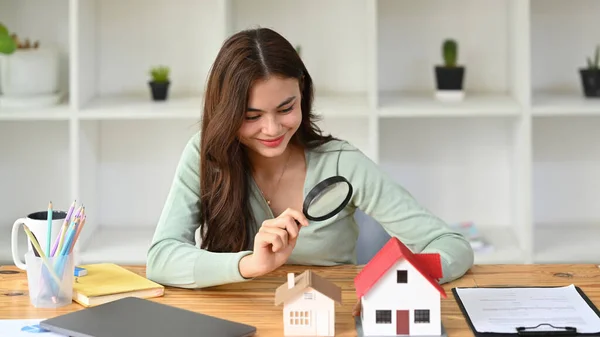 Smiling female appraiser looking at house model through magnifying glass. Real estate appraisal and house search concept.