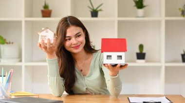 Happy young woman holding house models and smiling at camera. Real estate, mortgage loan and home insurance concept.