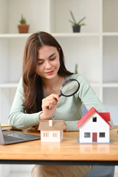 Female appraiser looking at house model through magnifying glass. Real estate appraisal and house search concept.