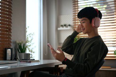 Smiling young man enjoy favorite songs on headphones and using mobile phone, spending leisure time at home in autumn day.