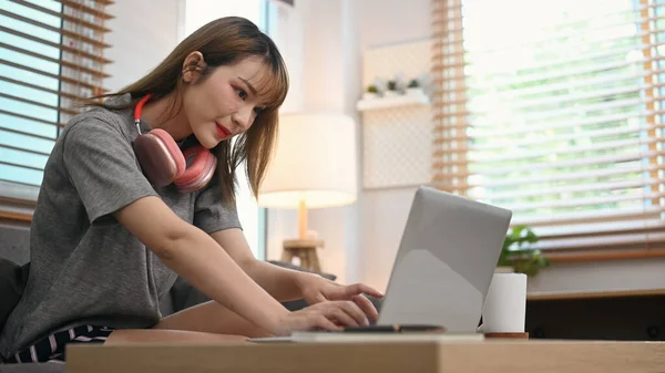 Beautiful young woman with wireless headphone using laptop on couch in comfortable living room.