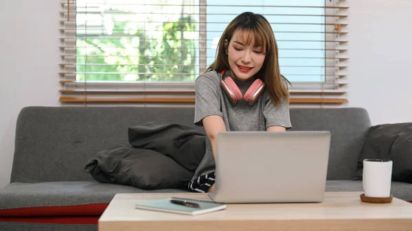 Pretty young woman with wireless headphone using laptop on comfortable couch. Entertainment and leisure activity concept.