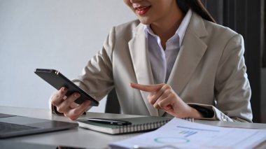 Smiling asian woman in business suit using mobile phone at her office desk.