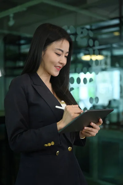 Smiling asian female team leader standing in corporate office and using digital tablet.