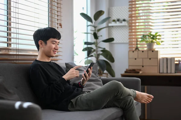 Relaxed young man in sweater using mobile phone, spending leisure time at home in winter or autumn weekend.